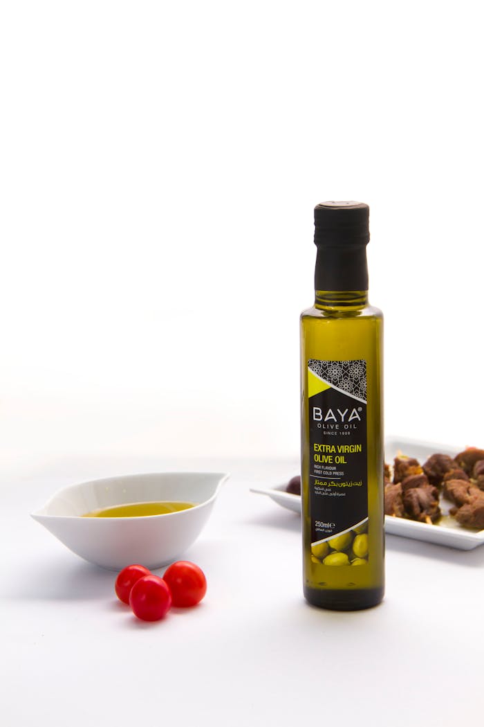 Extra virgin olive oil bottle with ceramic plate, cherry tomatoes, and grilled meat on white surface.